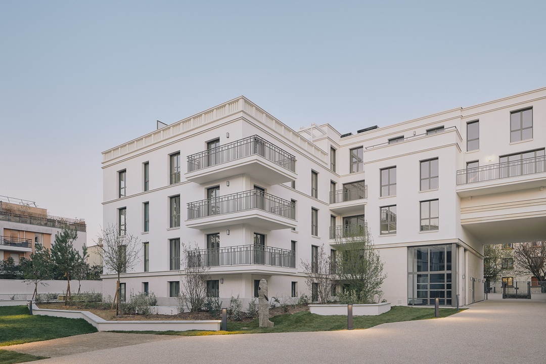 Résidence de logements contemporains aux façades blanches et balcons métalliques, organisée autour d’une cour paysagée avec cheminements piétons, arbres et espaces verts, photographiée en lumière douce de fin de journée.